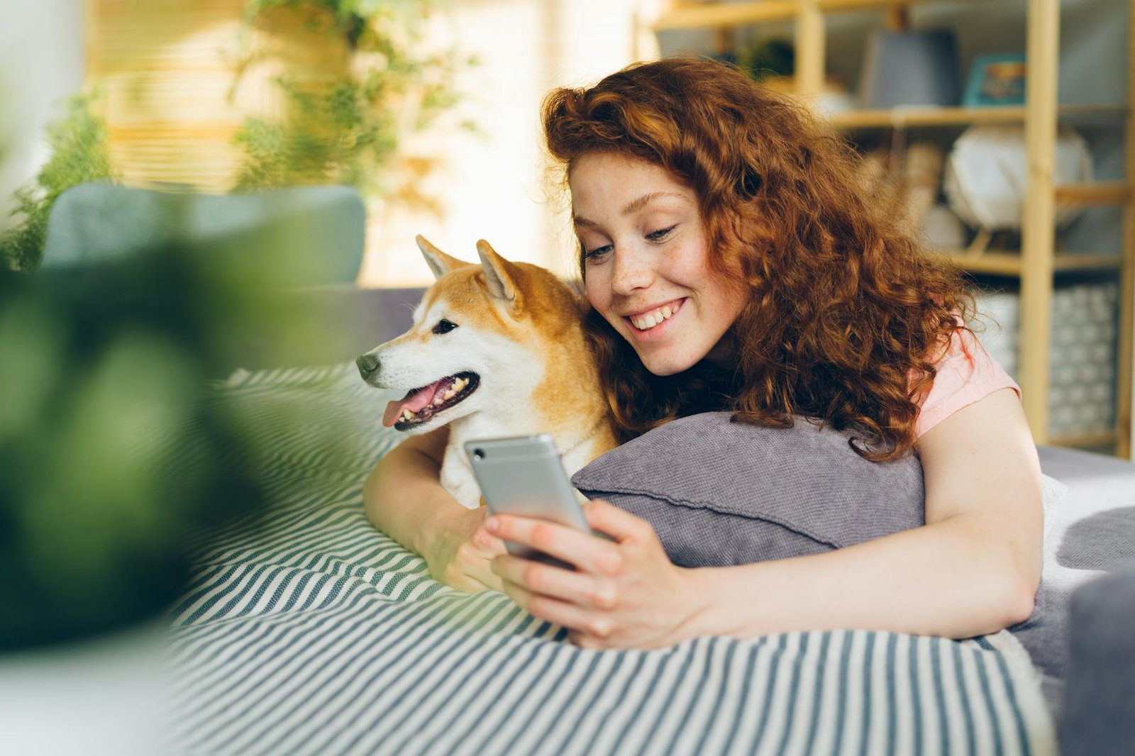 A smiling woman lying on a couch with her Shiba Inu dog and using her phone